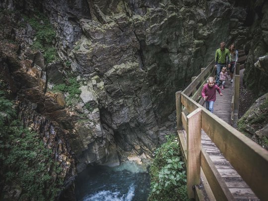 Dall’hotel a Colle Isarco a 3 stelle verso l’avventura Famiglia cammina su passerella in legno lungo una gola rocciosa con acqua