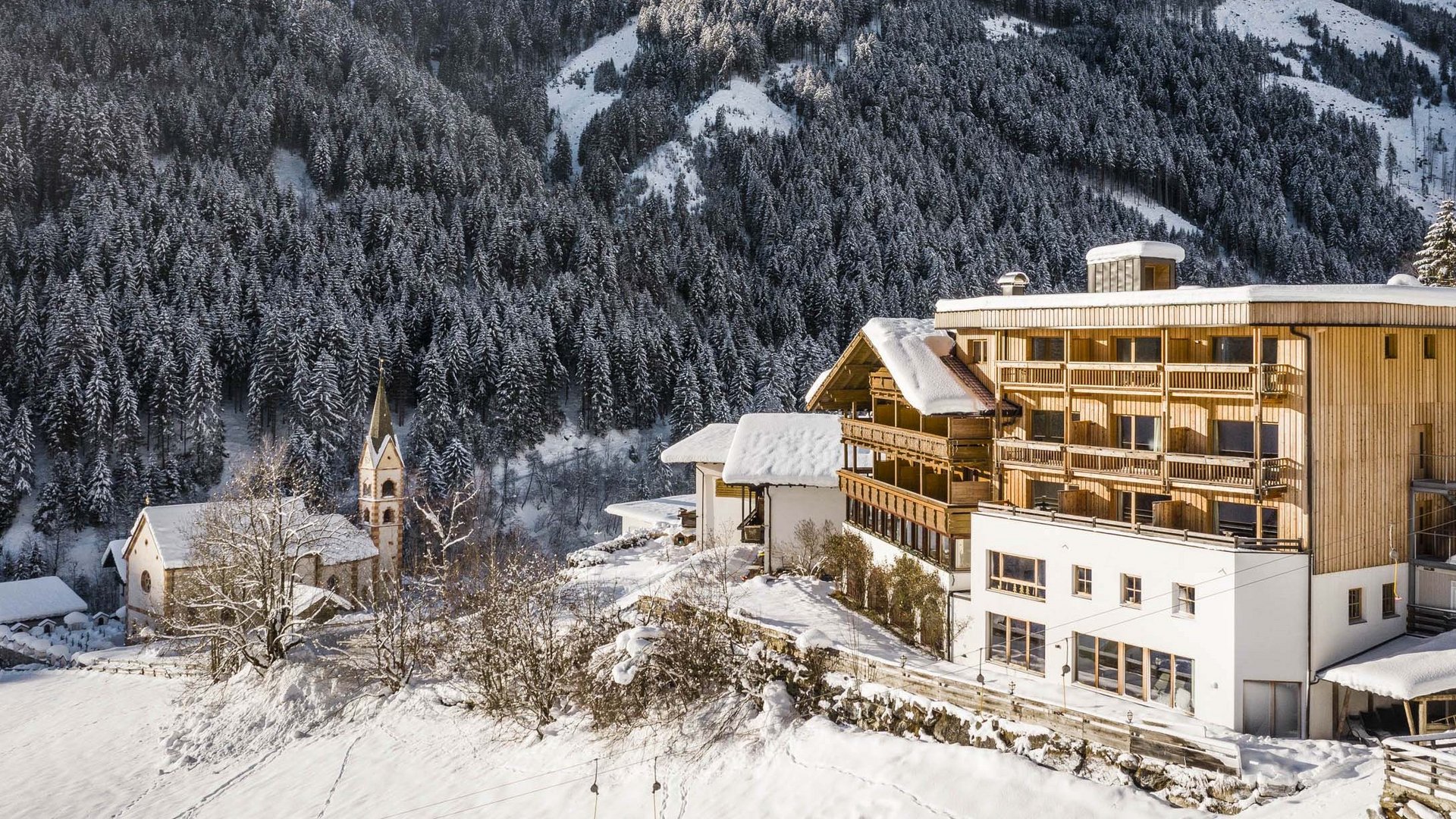 Family hotel in Sterzing: hoi, hello, and welcome! Snow-covered village houses and church below forested mountain peaks