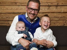 Familienhotel in Südtirol: Hotel Panorama Vater mit zwei lächelnden Kindern auf der Couch vor Holzwand