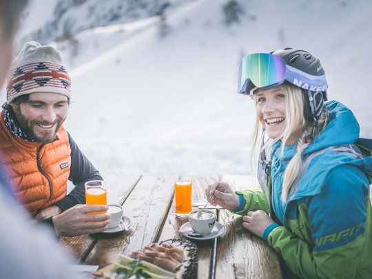 Skifahren in Ladurns Zwei Personen trinken Kaffee und Saft an einem verschneiten Außenholzttisch