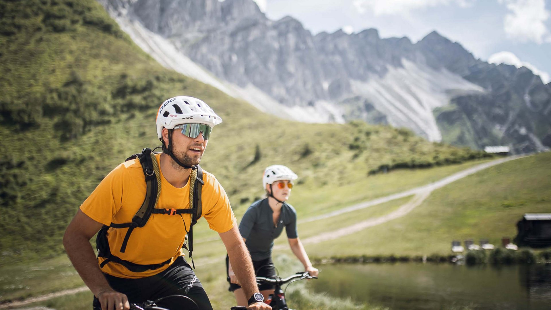 Lo sport a Colle Isarco vi coinvolgerà Due ciclisti con caschi pedalano su un sentiero di montagna accanto a un lago