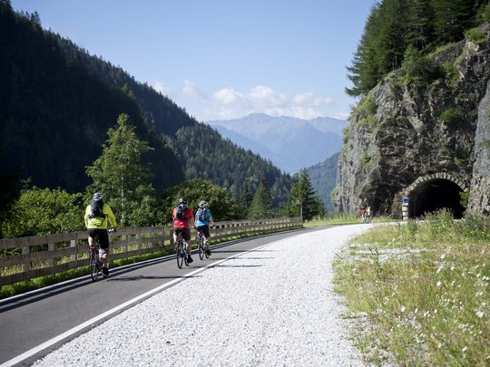Aktivhotel in Südtirol: Sommererlebnisse Radfahrer fahren auf einer Bergstraße Richtung Tunnel mit bewaldeten Hügeln im Hintergrund