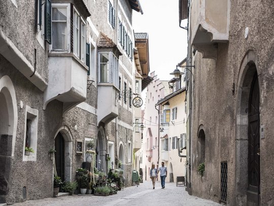 Hotel Panorama in Gossensass – Unterkunft zum Träumen Paar geht durch eine enge, historische Gasse mit alten Häusern und Blumen