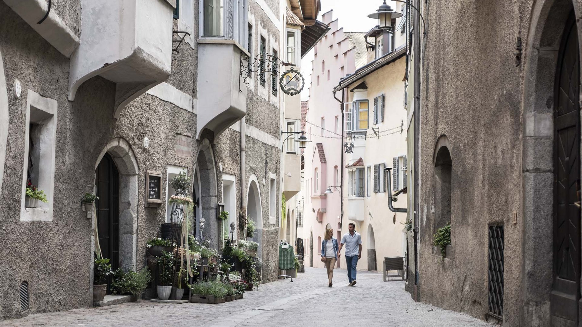 Dall’hotel a Colle Isarco a 3 stelle verso l’avventura Coppia che cammina in una stretta via storica con case antiche e fiori