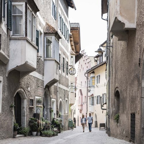 I nostri alloggi a Colle Isarco: una base per mille avventure Coppia che cammina in una stretta via storica con case antiche e fiori