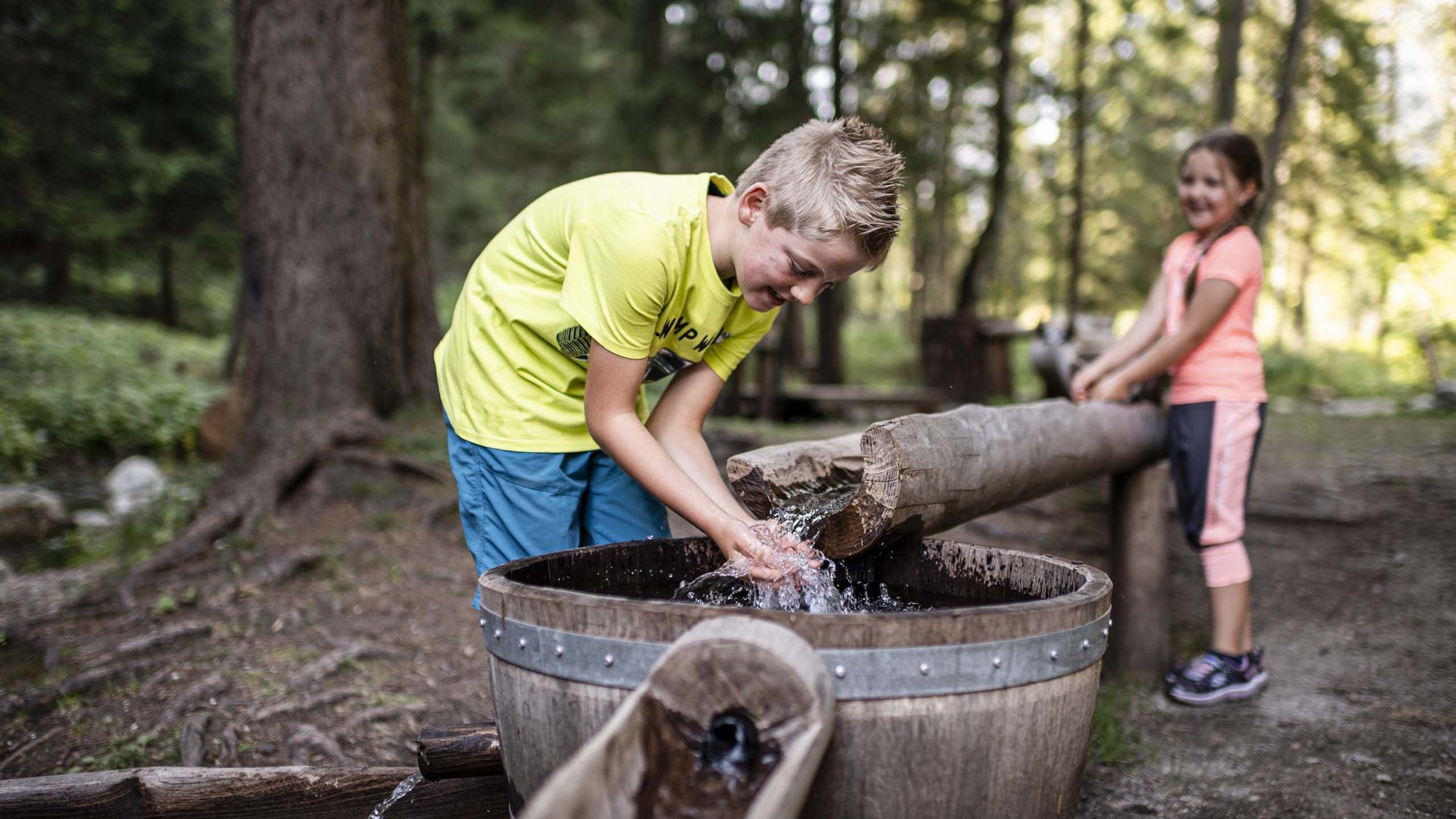 I nostri alloggi a Colle Isarco: una base per mille avventure Ragazzo e ragazza giocano con l'acqua a una fontana di legno nella foresta