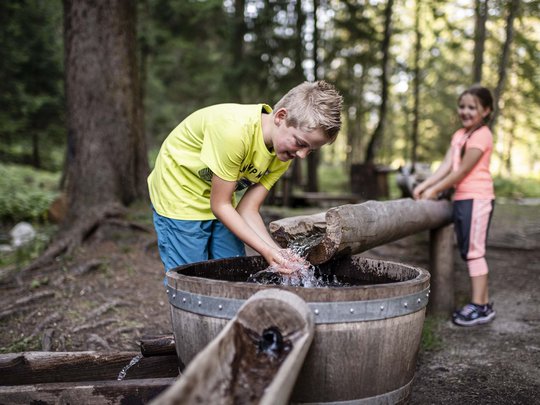Aktivhotel in Südtirol: Sommererlebnisse Junge und Mädchen spielen mit Wasser an einem traditionellen Holzbrunnen im Wald