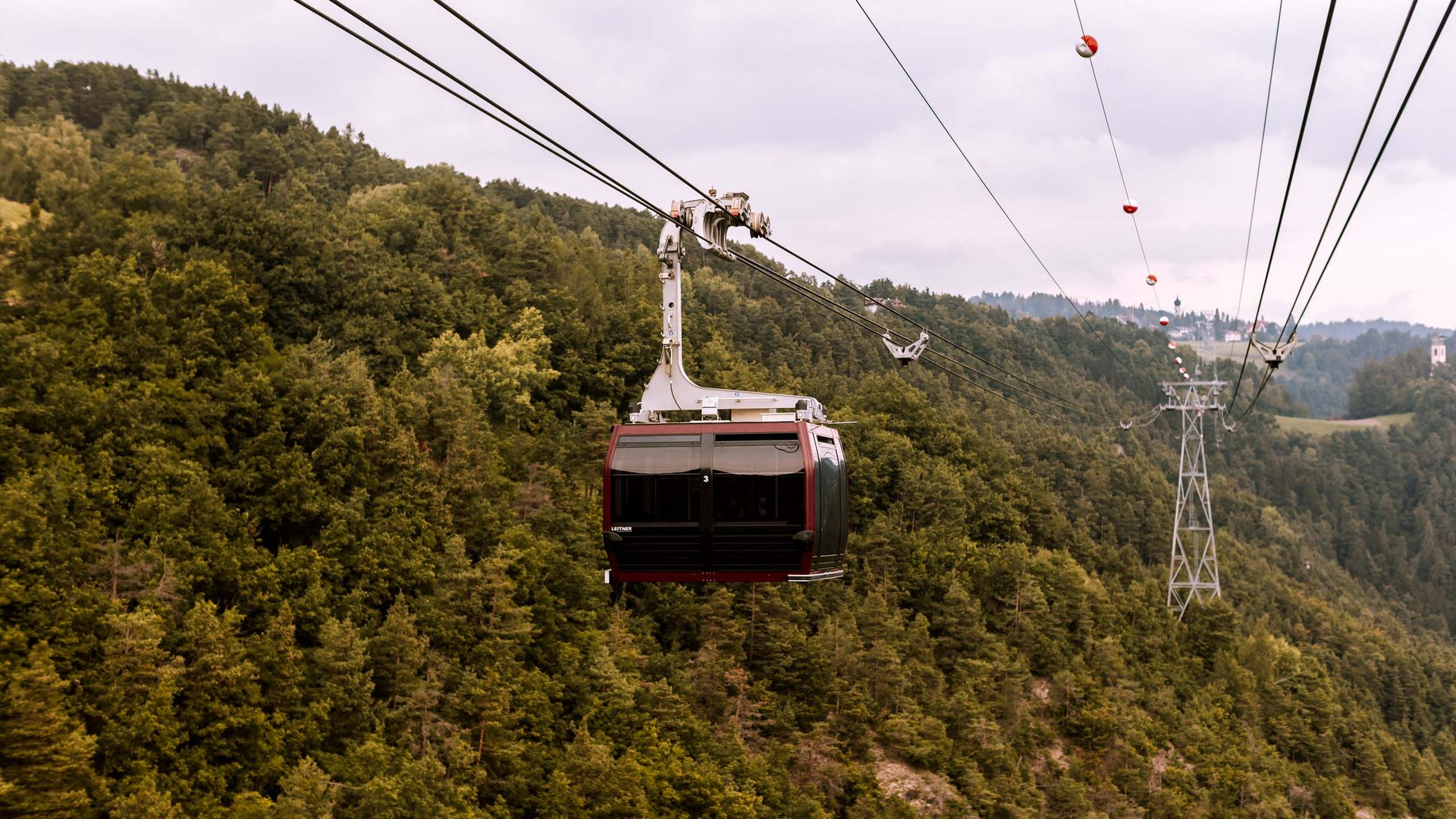Hotel mit Bergpanorama und GossensassCARD Seilbahn über bewaldeten Hügel mit grauem Himmel im Hintergrund