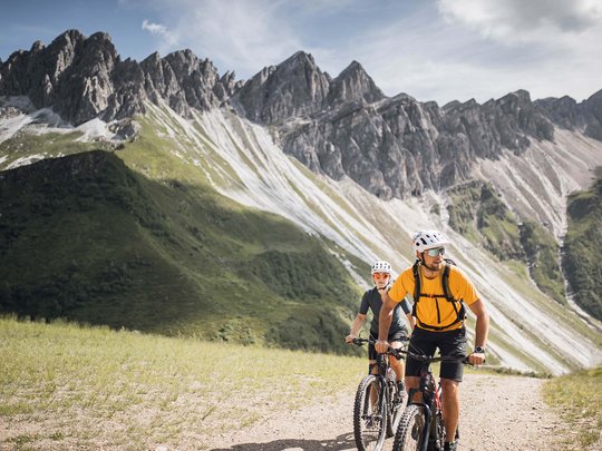 Lo sport a Colle Isarco vi coinvolgerà Due ciclisti su sentiero di montagna con vette frastagliate sullo sfondo